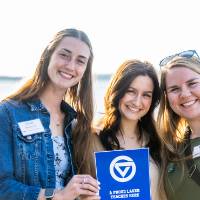 Group of 3 young women outside at event holding GVSU flag in front of them